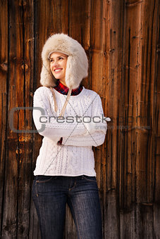 Woman in knitted sweater and furry hat standing near wood wall