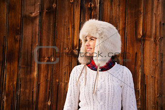 Happy woman in furry hat near rustic wood wall looking aside