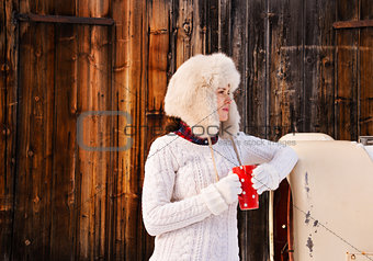 Woman in sweater and furry hat with cup near rustic wood wall
