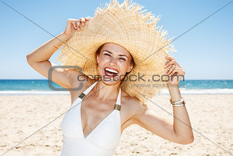 Smiling woman in swimsuit playing with big straw hat at beach