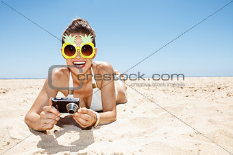 Smiling woman in pineapple glasses with photo camera at beach
