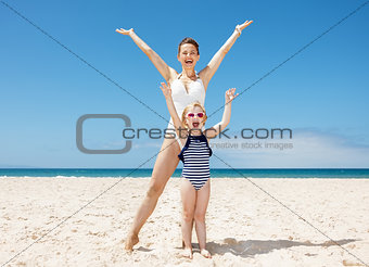 Happy mother and child in swimsuits at sandy beach rejoicing