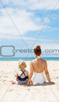 Seen from behind mother and child in swimsuits sitting at beach