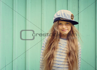 Cute little girl stands near a turquoise wall in sailor hat and smiling Space for text.