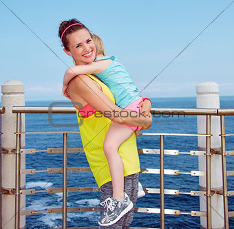 Smiling mother and child in fitness outfit hugging on embankment