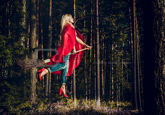Modern witch blond in jeans and red cape flying on a broom through the forest. Levitation. Halloween holiday.