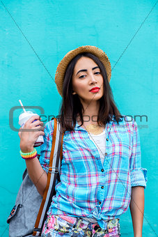 The girl in the city wearing shirt is holding a cocktail fresh  juice,  enjoying  backpack brunette, tanned, sensual make-up, recreation concept, travel, lifestyle. Summer day. Against the backdrop of  blue wall with red lipstick.