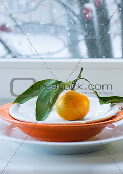 Tangerine with a branch and leaves on  plate against the background of  window  the snow