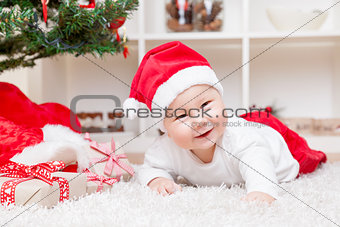 Cute baby in a Santa hat next to Christmas tree with presents
