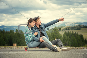 The guy with girl sit on a longboard. Man shows his hand forward. Road in the mountains.