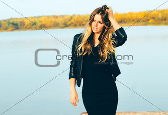 Young beautiful blonde woman in autumn park with lake in dark leather jacket smile  perfect teeth during sunset.