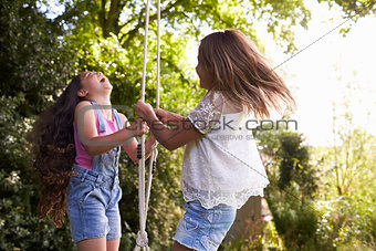 Two Girls Playing Together On Tire Swing In Garden