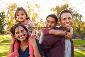 Mixed race parents carry their kids piggyback in a park