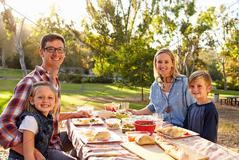 Young family at a picnic in a table in a park look to camera