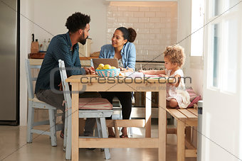 Girl Paints At Kitchen Table As Parents Look At Laptop