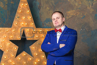 Happy elegant man in blue suit is standing with crossed hands at wall background.