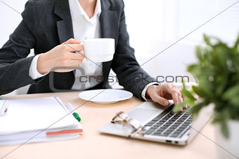 Close up of business woman hands with a cup of coffee is sitting at the table and typing on a laptop computer in the white colored office