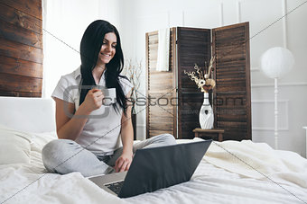 Relaxing on the bed. Beautiful woman enjoying a cup of coffee and using her laptop