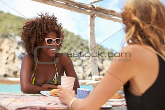 Two female friends sit talking at a table by the sea, Ibiza