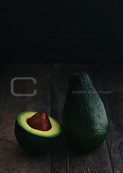 Two avocados, one of which cut in half on a dark, wooden backgro