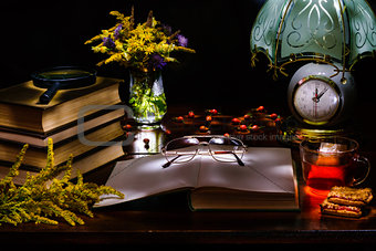 Still life of a pile of books,glasses,magnifier,vase with flowers,tea and biscuits,a lamp with a clock.Illuminated by the flashlight.