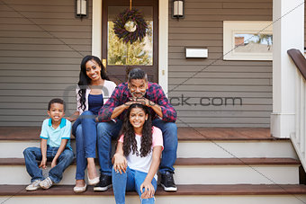 Family With Children Sit On Steps Leading Up To Porch Of Home