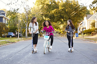 Three pre-teen girls riding in street on scooters and a bike