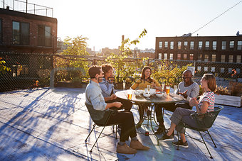Five friends sit talking at a table on a New York rooftop