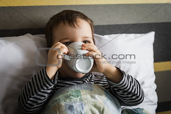 unhealthy little boy drinking cup of medicament lying on bed