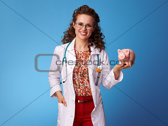 happy pediatrician doctor holding piggy bank on blue