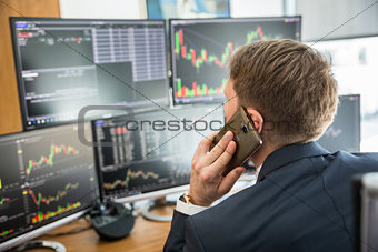 Over the shoulder view of stock broker trading online, talking on mobile phone.