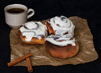 Buns with cinnamon and coffee on parchment paper and a dark background.