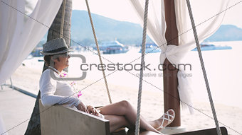 Happy woman in aged in hat and sunglasses resting on a swing on a tropical beach during summer vacation